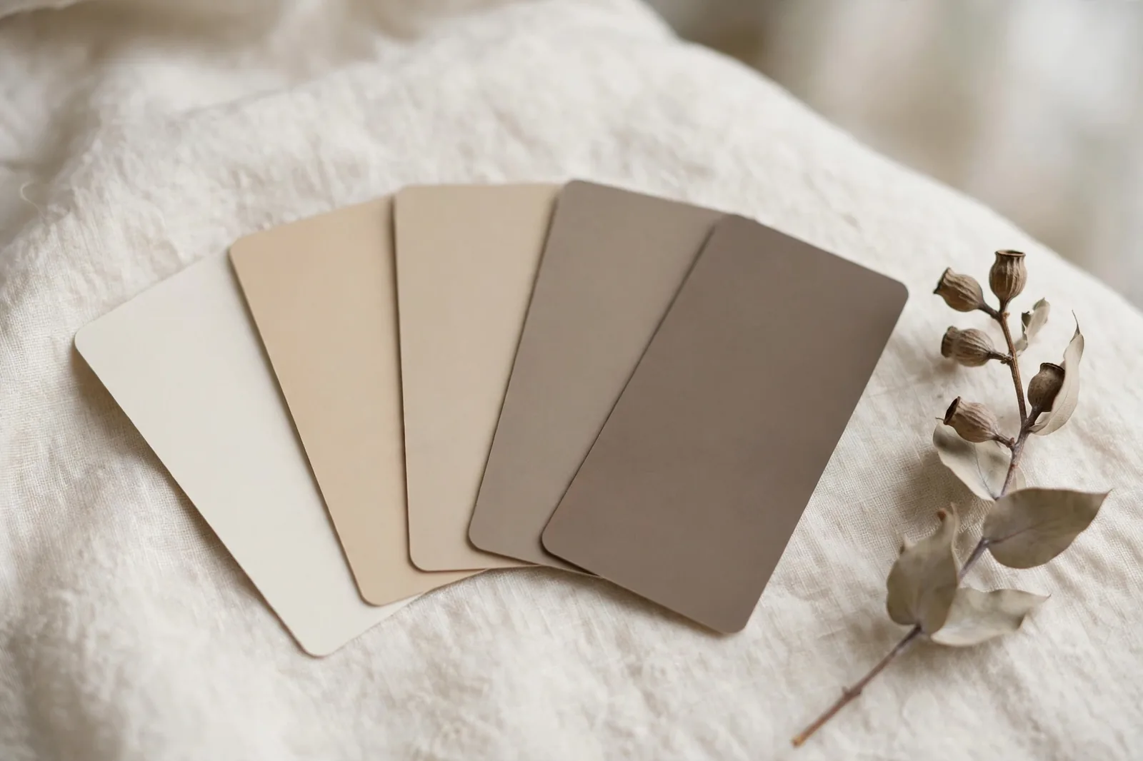 Five paint swatch cards in graduated warm neutral tones fanned on ivory linen